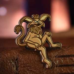 May include: A gold-colored enamel pin featuring a cartoon creature with a long tail, large ears, and a mischievous expression. The creature is sitting with its legs crossed and has a long, pointed tail.