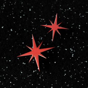 May include: Two bright red star-shaped pins on a black background speckled with white dots. The pins have a glossy finish and a seven-pointed design, evoking a celestial or space theme. The pins are likely accessories.