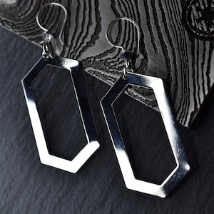 May include: A pair of silver geometric earrings with a hexagonal shape. The earrings are hanging from a textured surface.
