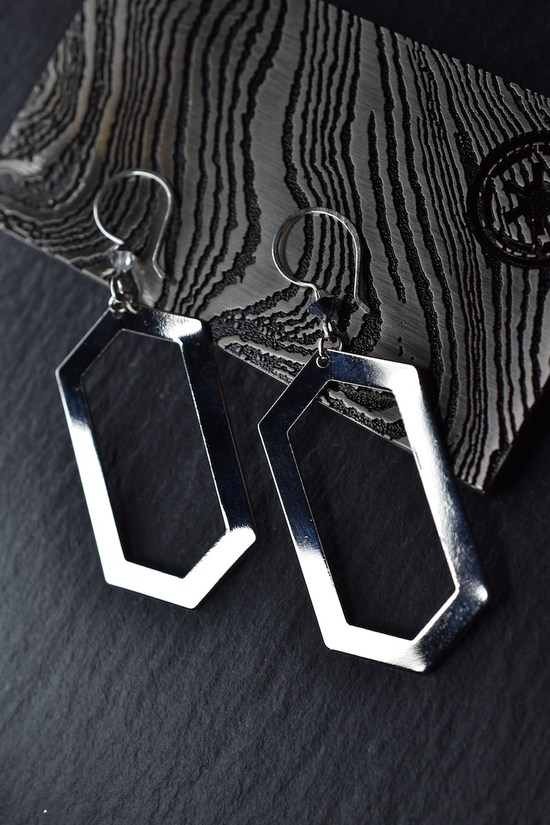 May include: A pair of silver geometric earrings with a hexagonal shape. The earrings are hanging from a textured surface.