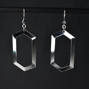 May include: A pair of silver geometric earrings with a hexagonal shape. The earrings are hanging from a black stand.