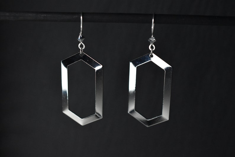 May include: A pair of silver geometric earrings with a hexagonal shape. The earrings are hanging from a black stand.