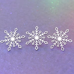 May include: Three white snowflake ornaments with intricate cut-out designs are arranged on a purple and iridescent background. The snowflakes have a symmetrical, six-pointed star shape, perfect for winter or holiday decor.