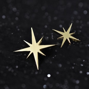 May include: Two gold-toned star-shaped metal pins with eight pointed rays of varying lengths. The larger star is positioned to the left, while the smaller star is placed to the right. Both stars feature a smooth, reflective, polished surface.