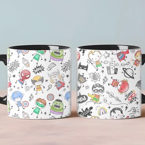 May include: Two ceramic mugs with black handles and rims. The mugs feature a white background with a repeating pattern of cartoon superheroes, speech bubbles, and action words. The mugs are on a light pink surface.