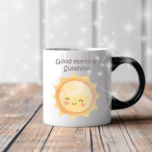 May include: White ceramic mug with a black handle featuring the text "Good morning Sunshine" and a smiling sun illustration. The sun has a yellow face with a happy expression. The mug is on a wooden surface.