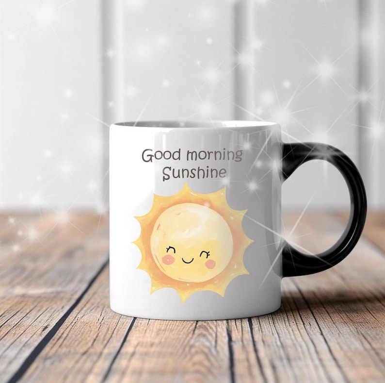 May include: White ceramic mug with a black handle featuring the text "Good morning Sunshine" and a smiling sun illustration. The sun has a yellow face with a happy expression. The mug is on a wooden surface.