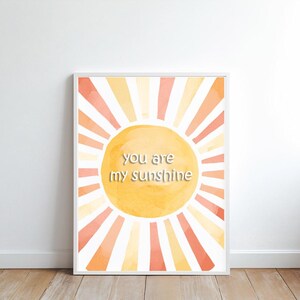 May include: Framed art print featuring a watercolor sun design with orange, yellow, and peach hues. The sunburst design has the text "you are my sunshine" in the center. The artwork is displayed on a white wall.