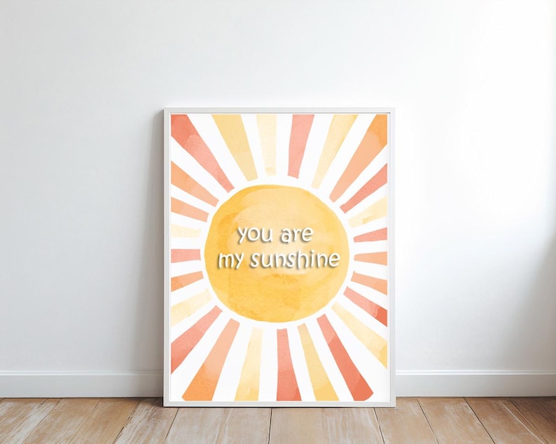 May include: Framed art print featuring a watercolor sun design with orange, yellow, and peach hues. The sunburst design has the text "you are my sunshine" in the center. The artwork is displayed on a white wall.