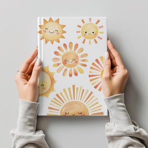 May include: A white journal featuring a repeating pattern of smiling sun illustrations in shades of yellow and orange. The journal is held by a person wearing a gray long-sleeved shirt.
