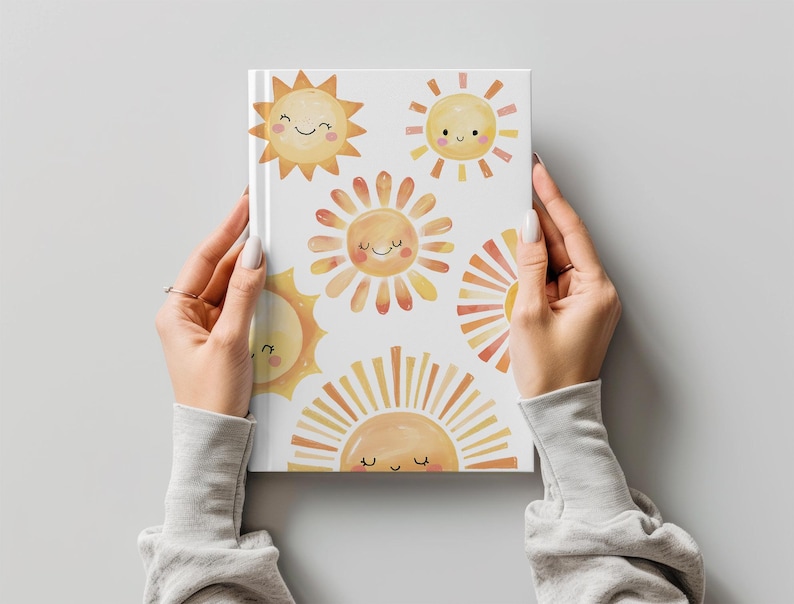 May include: A white journal featuring a repeating pattern of smiling sun illustrations in shades of yellow and orange. The journal is held by a person wearing a gray long-sleeved shirt.