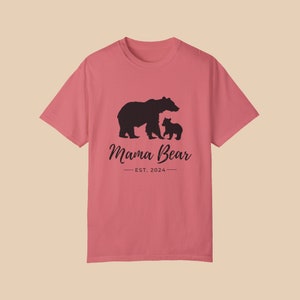 A 'Watermelon' color t-shirt with a black silhouette of a mama bear and a baby bear. The text 'Mama Bear' is written below the bears in a cursive font, and the text 'EST. 2024' is written below that in a sans serif font.