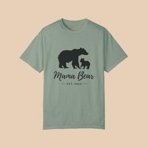 A 'bay' green color t-shirt with a black silhouette of a mama bear and a baby bear. The text 'Mama Bear' is written below the bears in a cursive font, and the text 'EST. 2024' is written below that in a sans serif font.