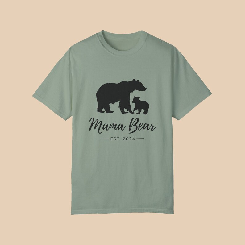 A 'bay' green color t-shirt with a black silhouette of a mama bear and a baby bear. The text 'Mama Bear' is written below the bears in a cursive font, and the text 'EST. 2024' is written below that in a sans serif font.