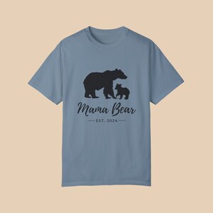 An 'ice blue' color t-shirt with a black silhouette of a mama bear and a baby bear. The text 'Mama Bear' is written below the bears in a cursive font, and the text 'EST. 2024' is written below that in a sans serif font.