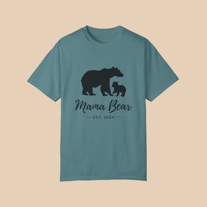 A 'blue spruce'' color t-shirt with a black silhouette of a mama bear and a baby bear. The text 'Mama Bear' is written below the bears in a cursive font, and the text 'EST. 2024' is written below that in a sans serif font.