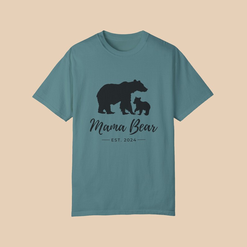A 'blue spruce'' color t-shirt with a black silhouette of a mama bear and a baby bear. The text 'Mama Bear' is written below the bears in a cursive font, and the text 'EST. 2024' is written below that in a sans serif font.