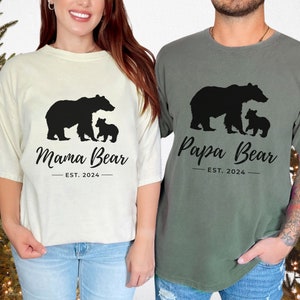 Two t-shirts, one white and one green, with a bear family silhouette design. The white t-shirt has the text 'Mama Bear - EST. 2024' and the green t-shirt has the text 'Papa Bear - EST. 2024'.