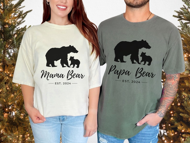 Two t-shirts, one white and one green, with a bear family silhouette design. The white t-shirt has the text 'Mama Bear - EST. 2024' and the green t-shirt has the text 'Papa Bear - EST. 2024'.