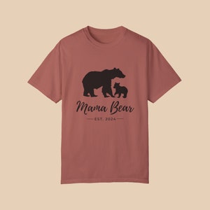 A 'cumin' color t-shirt with a black silhouette of a mama bear and a baby bear. The text 'Mama Bear' is written below the bears in a cursive font, and the text 'EST. 2024' is written below that in a sans serif font.