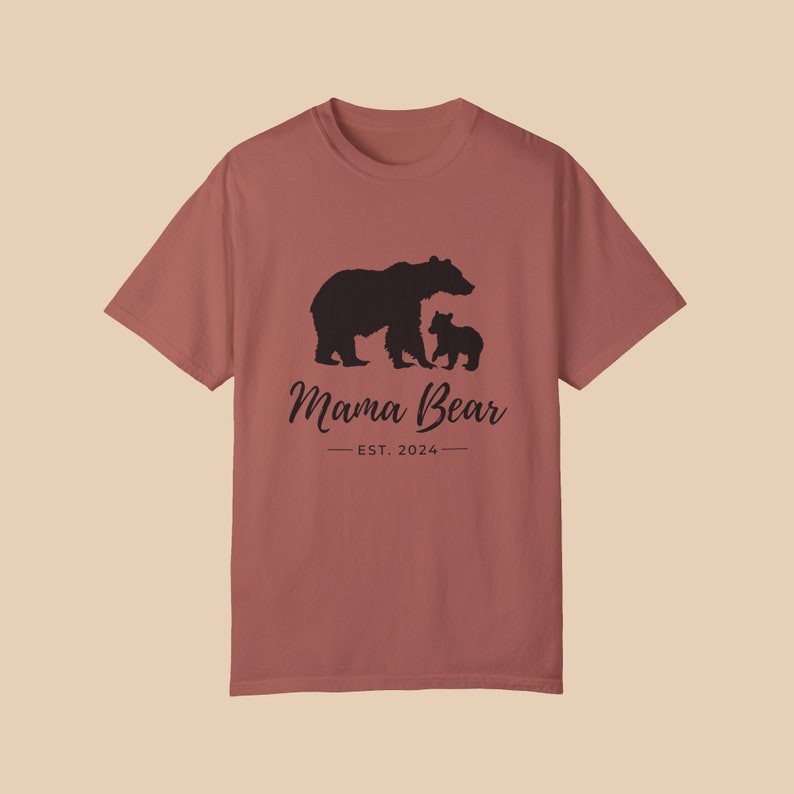 A 'cumin' color t-shirt with a black silhouette of a mama bear and a baby bear. The text 'Mama Bear' is written below the bears in a cursive font, and the text 'EST. 2024' is written below that in a sans serif font.