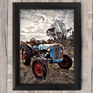 May include: Framed print of a vintage blue tractor with red wheels, set against a cloudy sky and dry field. The tractor shows signs of age with rust, adding to its rustic charm. The artwork is framed in a black wooden frame.