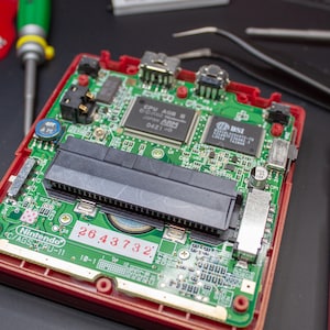 Könnte beinhalten: Nahaufnahme einer grünen Leiterplatte mit einem schwarzen Kartensteckplatz. Die Platine hat mehrere Chips und Komponenten, darunter einen CPU AGB-Chip mit dem Text "JAPAN ARM 0421-0". Die Platine ist mit "Nintendo C/AGS CRU-11 10-1" beschriftet und hat die Seriennummer "2643732".
