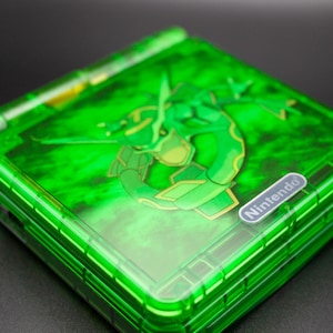 May include: A green Nintendo Game Boy Advance SP console with a translucent cover. The cover features a green dragon graphic and a small Nintendo logo. The console is a vibrant shade of green.