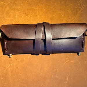 May include: A brown leather pencil case with a strap closure and brass accents.