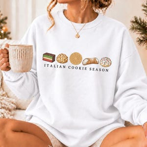 May include: A white crewneck sweatshirt featuring a graphic of five assorted Italian cookies and the text "ITALIAN COOKIE SEASON" printed in black. The design includes a layered cake, a round textured cookie, and a cannoli.