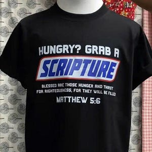 May include: Black t-shirt with the text "HUNGRY? GRAB A SCRIPTURE" in white and blue. Below the text is "BLESSED ARE THOSE HUNGER AND THIRST FOR RIGHTEOUSNESS, FOR THEY WILL BE FILLED MATTHEW 5:6".