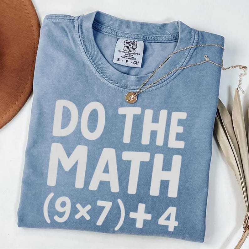 May include: A light blue t-shirt with the phrase "DO THE MATH" in large white letters. Below the phrase is the equation (9x7)+4. A gold necklace with a small pendant rests on the shirt. The shirt is folded and has a tag that says "Comfort Colors".