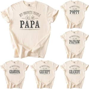 May include: Cream-colored t-shirts with various text designs. One shirt says "MY FAVORITE PEOPLE CALL ME PAPA." Other shirts say "POPPY," "PAPAW," "GRANDPA," "GRUMPY," and "GRANDY."