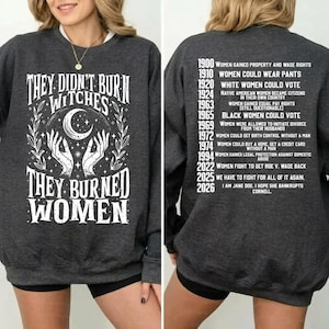 May include: A dark gray crewneck sweatshirt featuring white graphic text on the front reading "THEY DIDN'T BURN WITCHES THEY BURNED WOMEN" with a crescent moon and hands. The back displays a chronological list of historical dates and events.