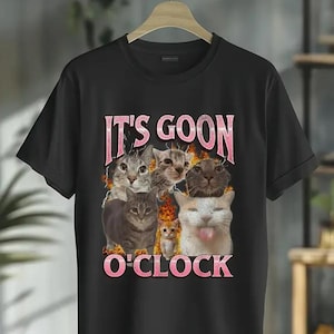 May include: Black t-shirt featuring a graphic of several cats with the text "IT'S GOON O'CLOCK" in pink letters. The cats have various expressions, and the design has a playful, humorous theme.