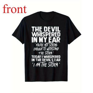 May include: Black t-shirt with white text that reads "THE DEVIL WHISPERED IN MY EAR" followed by a quote: "YOU'RE NOT STRONG ENOUGH TO WITHSTAND THE STORM" and "I AM THE STORM". The shirt has a crew neck and short sleeves.