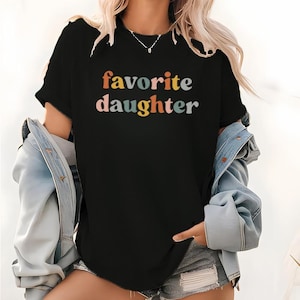 May include: Black t-shirt with the words "favorite daughter" in colorful, retro-style lettering. The shirt is short-sleeved and appears to be made of a soft material. The shirt is being worn with a denim jacket.
