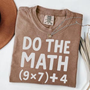 May include: A brown t-shirt with the phrase "DO THE MATH (9x7)+4" printed in white. The shirt has a crew neck and short sleeves. A silver necklace with a pendant is draped across the shirt.