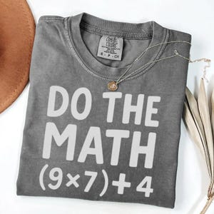 May include: A gray Comfort Colors t-shirt with the phrase "DO THE MATH (9x7)+4" printed in white. A gold-colored necklace with a pendant is draped over the shirt. The shirt is folded on a white surface.
