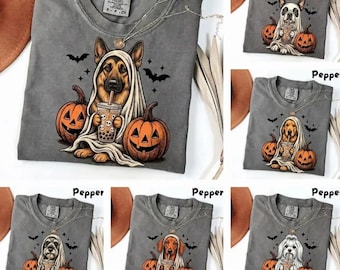 May include: A charcoal gray t-shirt featuring a graphic of a German Shepherd dog wearing a white ghost sheet, holding a boba tea, and surrounded by two orange jack-o'-lantern pumpkins and black bats. The shirt has a crew neck and a tag.