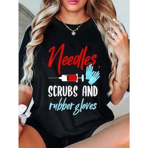 May include: Black t-shirt with the words "Needles, Scrubs and rubber gloves" in red, white, and blue. The design includes a syringe and blue medical gloves. A medical-themed graphic tee.