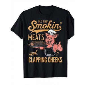 May include: Black t-shirt with a graphic featuring a smiling pig chef holding a platter of meat and giving a thumbs up. The text reads "Old Row Smokin' Meats and Clapping Cheeks" in a vintage style.