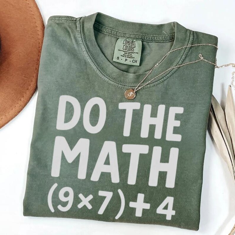 May include: Olive green t-shirt with the phrase "DO THE MATH" in white block letters, followed by the equation (9x7)+4. A gold necklace with a pendant rests on the shirt. The shirt is folded.