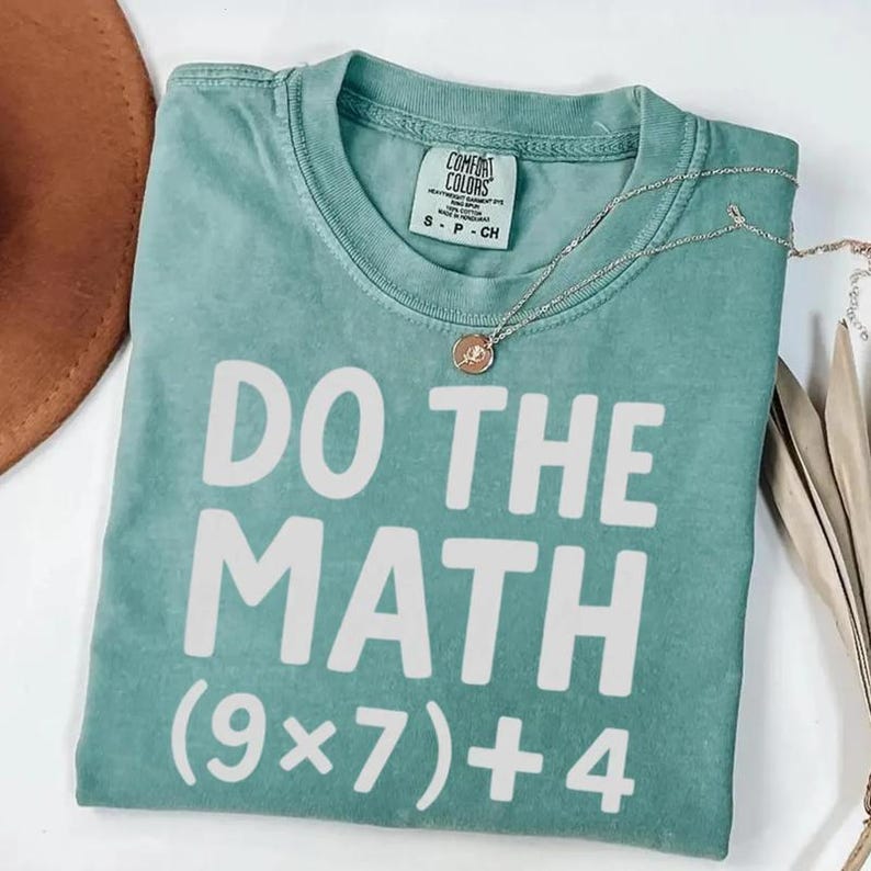 May include: A teal Comfort Colors t-shirt with the phrase "DO THE MATH" in large white block letters. Below the phrase is the equation (9x7)+4. A silver necklace with a pendant rests on the shirt.