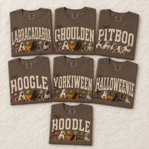 May include: Seven brown crewneck t-shirts featuring various dog breeds dressed in Halloween costumes. Each shirt displays a unique punny text including Labracadabra, Ghoulden, Pitboo, Boogle, Yorkiween, Halloweenie, and Boodle with polka dot lettering.