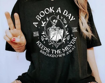 May include: Black t-shirt with a white graphic featuring a skeleton reading a book, surrounded by butterflies and the text "A Book A Day Keeps The Mental Breakdown Away." Additional text reads "Read More" and "Cry Less."
