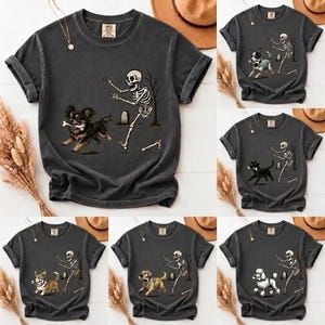 May include: A charcoal gray short-sleeve t-shirt featuring a graphic print of a skeleton chasing various dog breeds, including a chihuahua, a black dog, a corgi, a golden retriever, and a poodle, with a tombstone and a bone in the background.