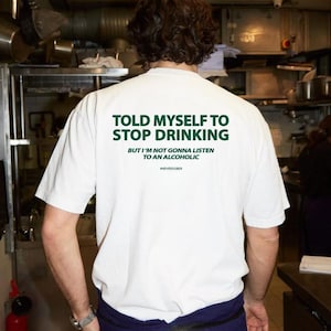 May include: White t-shirt with green text on the back that reads "TOLD MYSELF TO STOP DRINKING" above "BUT I'M NOT GONNA LISTEN TO AN ALCOHOLIC" and the hashtag "#NEVERSOBER". The shirt is being worn by a person.