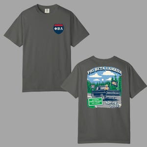 May include: Dark gray t-shirt with a small shield logo on the front and a graphic on the back. The back graphic features a vintage truck, trees, and the text "PHI BETA LAMBDA" and "Spring 2025".