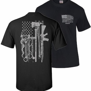 May include: Two black t-shirts. One features an American flag design with the text "NO ONE FIGHTS ALONE." The other shirt displays a flag design with symbols representing dispatch, fire, nurse, police, and military.